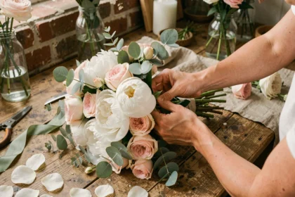 Mains de fleuriste composant un bouquet de mariée avec des pivoines blanches, roses garden et eucalyptus sur une table en bois rustique