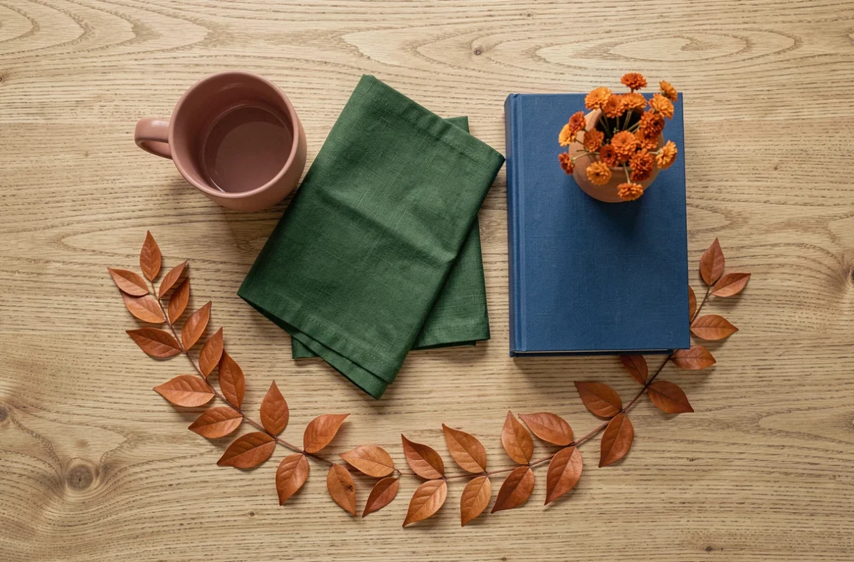 Flat lay de couleurs harmonieuses avec tasse rose, serviette verte, livre bleu cobalt et fleurs orange séchées sur une table en bois de chêne